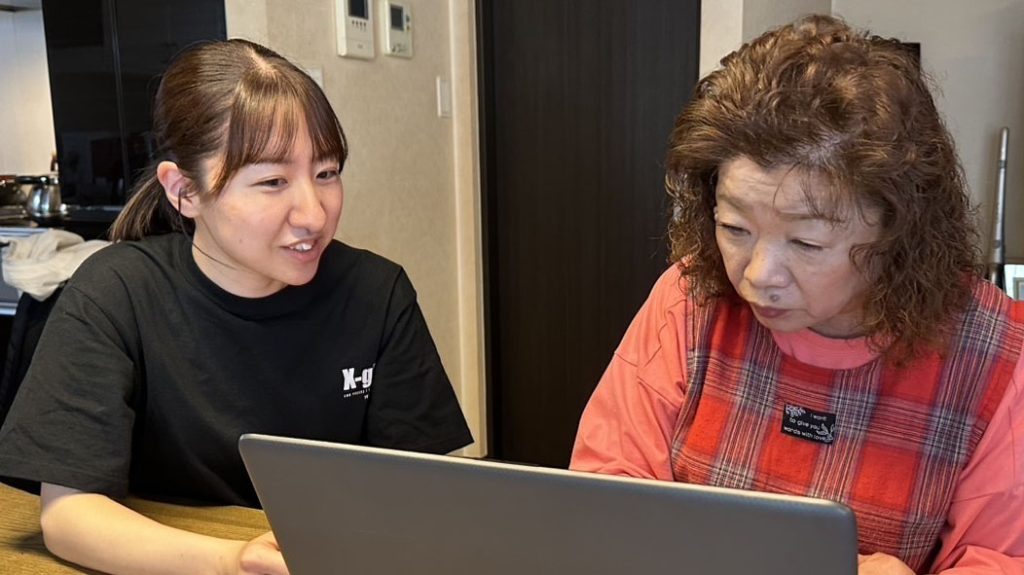 高齢者の女性に大学生がパソコンの使い方を教えている画像。